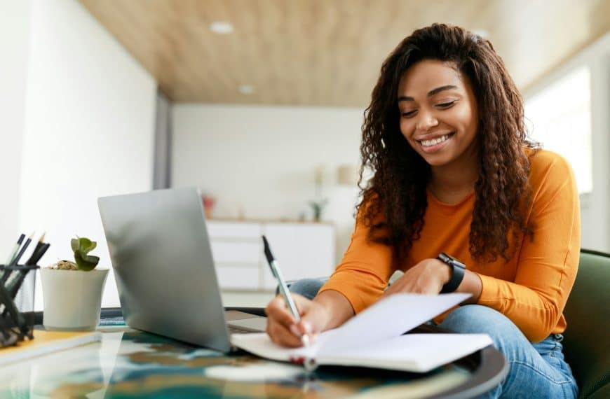 Black woman sitting at desk, using computer writing in notebook