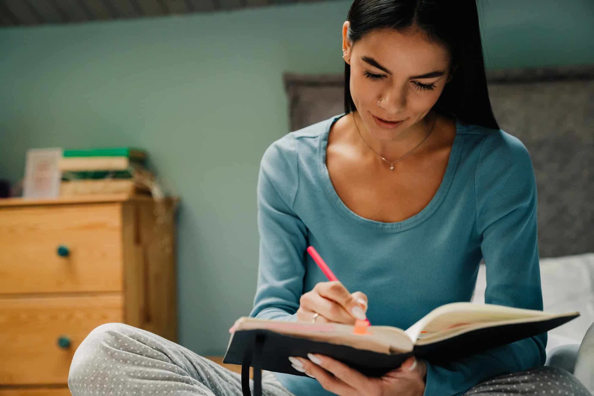 Young hispanic woman writing down notes at notebook in bed at home